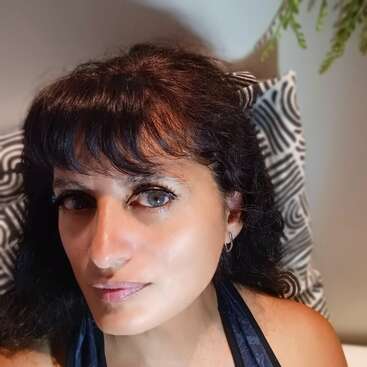 Uma mulher com cabelos escuros e franja olha para a câmera, sentada contra um travesseiro estampado. A iluminação destaca sua pele clara e seus olhos expressivos. Planta visível.