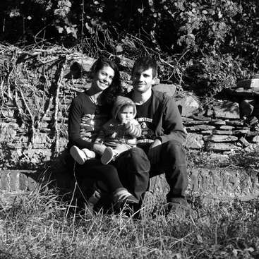 La imagen muestra a una familia de tres miembros sentados en un muro de piedra, con la madre sosteniendo a un bebé y el padre a su lado, todos sonriendo a la cámara.