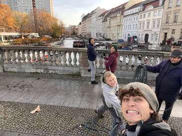 Vier lächelnde Menschen stehen auf einer Uferpromenade mit einer alten Brücke und einem Kanal hinter ihnen, umgeben von schönen historischen Gebäuden und herbstlich gefärbten Bäumen.