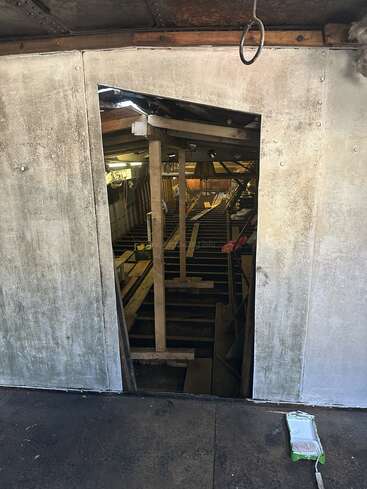 A roughly cut doorway in a weathered wall reveals the interior of a construction or renovation project, with exposed wood beams, construction materials, and bright lighting inside.