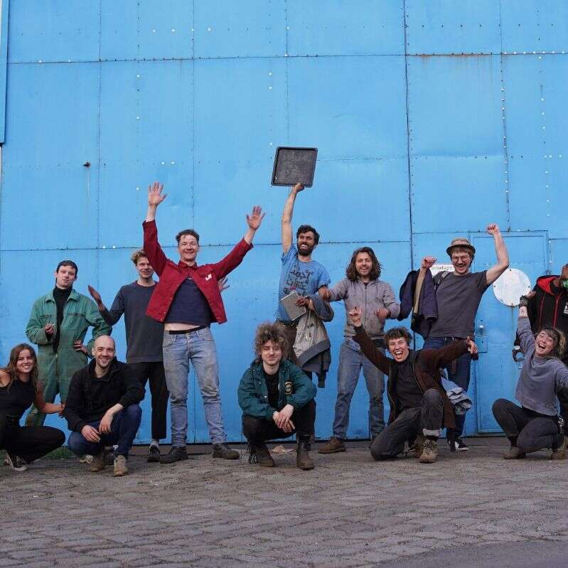 Das Bild zeigt eine Gruppe von 13 Personen, die für ein Foto vor einer großen blauen Wand posieren, wobei einige Personen ihre Arme zum Feiern heben.