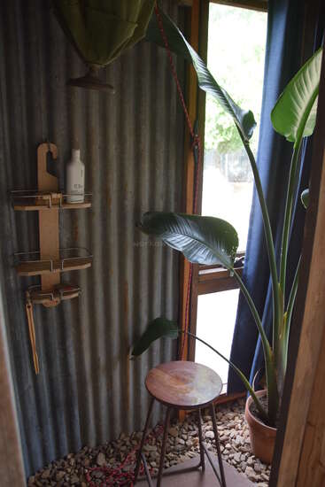 A rustic shower with corrugated metal walls, a wooden stool, indoor plant, pebbled floor, wall-mounted shelf holding toiletries, and sunlight streaming through a curtained window.
