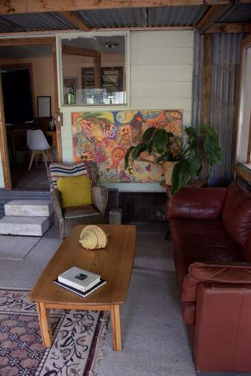 A cozy, rustic room with a red leather couch, wicker chair, colorful abstract painting, wooden table with books, potted plant, and bohemian decor details throughout.