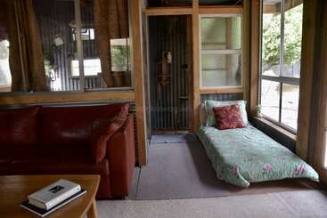 A cozy room contains a red couch, a coffee table with two books, a single bed with floral bedding, large windows, and rustic wooden walls.