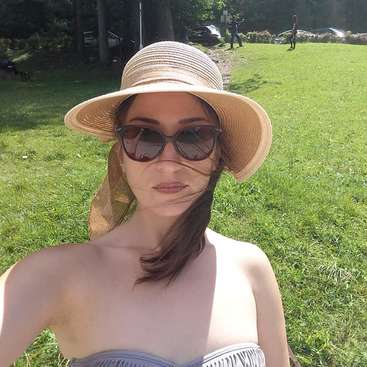 The image depicts a woman wearing a straw hat and sunglasses, posing for a selfie in a grassy field with trees and cars in the background on a sunny day.