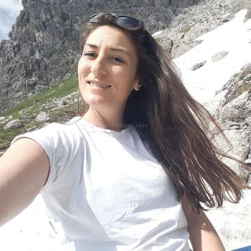 Eine Frau mit langen dunklen Haaren lächelt, während sie draußen ein Selfie macht. Sie trägt ein weißes T-Shirt und eine Sonnenbrille, hinter ihr sind felsige Berge und Schneeflecken zu sehen.