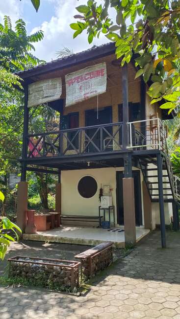A two-story house surrounded by lush greenery, with a “WELCOME” sign on the balcony, outdoor seating below, and a staircase leading to the upper floor.