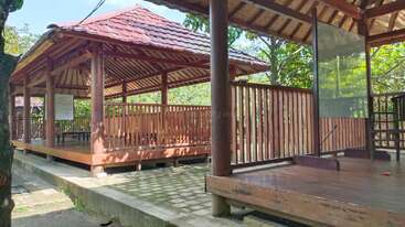 The image shows two traditional wooden gazebos with red tiled roofs, elevated floors, and open sides, surrounded by greenery and shaded by trees in a peaceful setting.