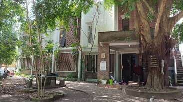 The image shows a shaded courtyard with large trees, circular stone benches, and a brick building featuring open windows, stairs, and people’s belongings at the entrance.