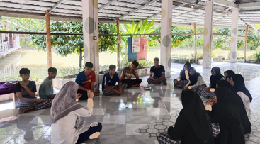 A group of young people sit in a circle on a tiled floor in an open, roofed area near a pond, engaged in a discussion.
