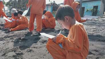 A group of children dressed in matching orange outfits sit on a sandy beach, focused on writing or drawing on white papers, enjoying an outdoor learning activity.