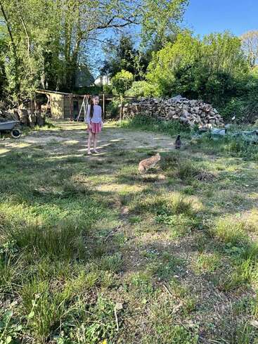 Una niña está descalza en un jardín cubierto de hierba, con un gato y una gallina cerca. Al fondo se ven árboles, leña apilada y un cobertizo.