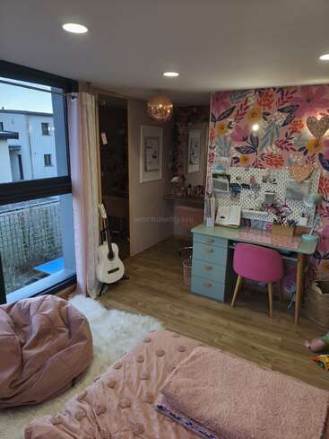 Esta acogedora habitación cuenta con una pared decorativa con motivos florales, un escritorio en tonos pastel, una silla rosa, un cómodo puf, ropa de cama suave, una guitarra, una gran ventana y un cálido suelo de madera.