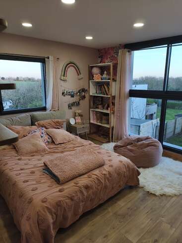 Una acogedora habitación con grandes ventanas, iluminación suave y decoración en tonos rosados. Cuenta con una cama, una estantería, un puf, una mullida alfombra, fotos y un tapiz con un arcoíris.