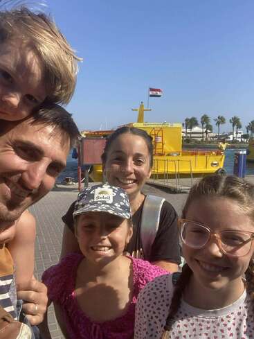 Une famille heureuse de cinq personnes pose pour un selfie en plein air par une journée ensoleillée, devant un bateau jaune et des palmiers au bord de l'eau.
