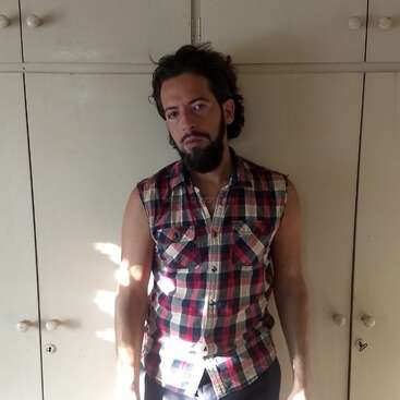 Ein Mann mit Bart und dunklen Haaren, der ein ärmelloses kariertes Hemd trägt, steht vor cremefarbenen Schränken und schaut nachdenklich, während das Sonnenlicht Schatten neben ihn wirft.