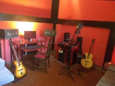 Ein gemütliches Musikstudio mit orangefarbenen Wänden, Computertisch, Lautsprechern, zwei Akustikgitarren, Mikrofonen, Stuhl und verschiedenen Musikgeräten, die für die Aufnahme und Produktion eingerichtet sind.