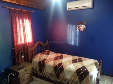 Ein gemütliches Schlafzimmer mit blauen Wänden, einem Einzelbett mit einer karierten Decke, einem hölzernen Nachttisch, roten Vorhängen, einer an der Wand befestigten Klimaanlage und einem kleinen Bild.