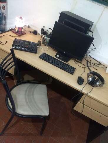 Ein Schreibtisch mit Computermonitor, Tastatur, Maus, Kopfhörer, Audiomixer, Lampe und einem Stuhl auf einem gefliesten Boden. Einige Kabel und elektronische Geräte sind sichtbar.