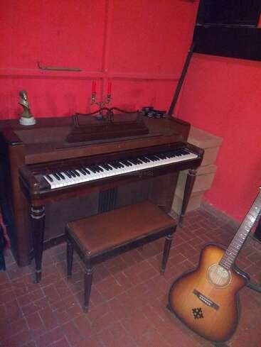 Das Bild zeigt ein Vintage-Klavier mit einer Bank, einen Kerzenleuchter, eine kleine Büste, gestapelte Kisten und eine Akustikgitarre vor rot gestrichenen Wänden. Gemütliche Atmosphäre.