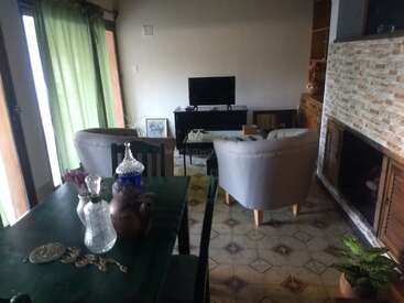 Ein gemütliches Wohnzimmer mit zwei grauen Sesseln, einem Fernseher, einem Kamin, grünen Vorhängen, einem Esstisch mit Dekoartikeln und dekorativen Bodenfliesen. Pflanzen in der Nähe.