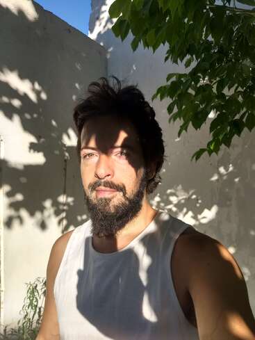 Un hombre barbudo con camiseta blanca de tirantes está de pie al aire libre, la luz del sol y las sombras de las hojas juegan con su rostro y el fondo, creando un efecto artístico y natural.