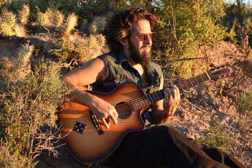 Ein bärtiger Mann mit Sonnenbrille sitzt in der Wüste zwischen Kakteen und Büschen, spielt auf einer Akustikgitarre und genießt die Musik und die Natur im goldenen Sonnenlicht.