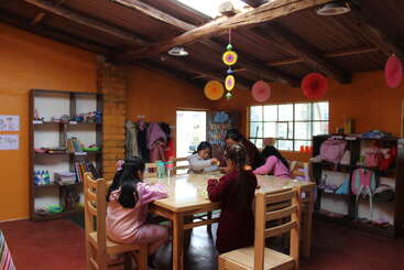 Five children sit around a wooden table in a colorful classroom, engaging in activities. The room has shelves, decorations, backpacks, and warm natural light.