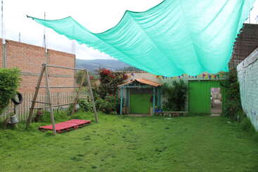 A grassy yard features a swing set, small wooden structure, and green tarp overhead. Bright flowers, colorful bunting, and a distant mountain backdrop add charm.