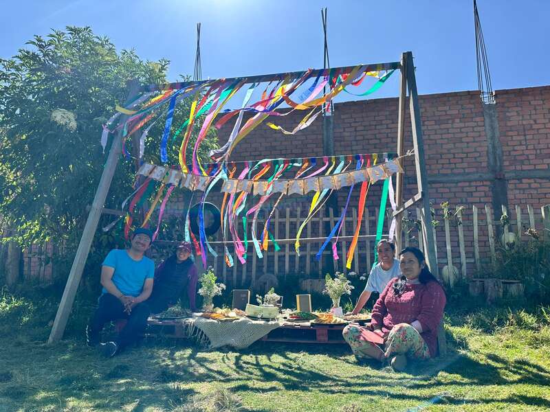 Four people sit on grass around a picnic table under a wooden frame decorated with colorful ribbons, enjoying a sunny outdoor gathering in a peaceful garden.
