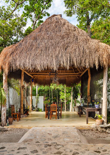 La imagen muestra una serena zona de comedor al aire libre, con una cabaña de techo de paja con mesa y sillas, rodeada de exuberante vegetación y árboles.