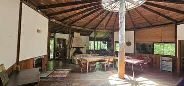 This spacious, rustic round room features a wooden ceiling, central support beam, charming fireplace, large windows, natural light, dining area, cozy sofas, and earthy decor throughout.