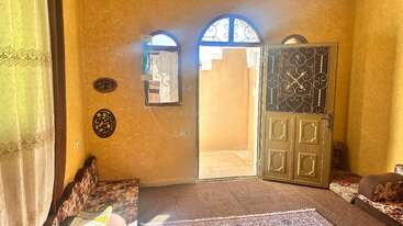 This image shows a cozy, sunlit room with yellow walls, patterned cushions, Islamic wall art, a carpet, and an open ornate door leading outside.