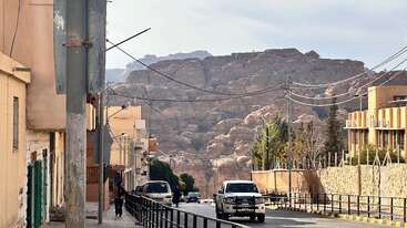A quiet street with a few cars and people, bordered by buildings and power lines. Majestic rocky mountains rise impressively in the background under a pale sky.