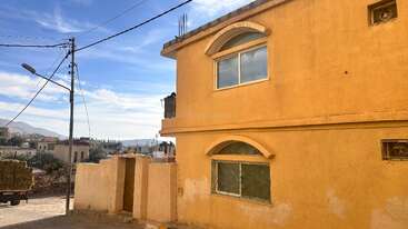 A two-story yellow building stands under a blue sky. Windows have decorative arches. Nearby, a wall and gate face a street with utility poles and distant mountains.