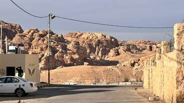 A quiet desert street ends at rugged, sunlit rocky hills. A white car, utility poles, and beige buildings stand under a clear sky, evoking calm isolation.