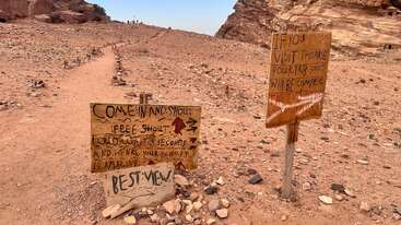 A desert trail leads between rocky hills. Two handmade wooden signs invite visitors to shout and hear echoes, promising the best view and unforgettable experience.