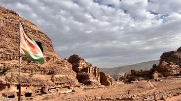 A Jordanian flag waves in the foreground, with magnificent rock-cut structures and rugged cliffs of Petra under a dramatic cloudy sky in the background. Stunning landscape.