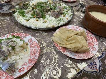 A traditional meal is served on a patterned tablecloth, featuring rice with lamb, flatbread, a creamy sauce, and garnished with herbs and nuts. Plates ready.