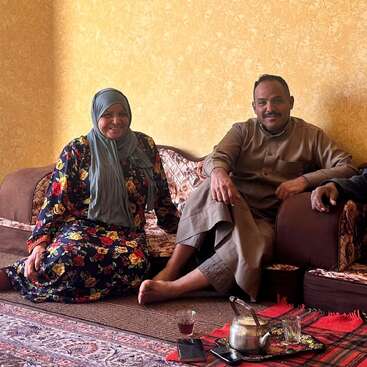 A smiling woman in a floral dress and headscarf sits beside a man in traditional clothing. They sit comfortably on a sofa, sharing tea together indoors.