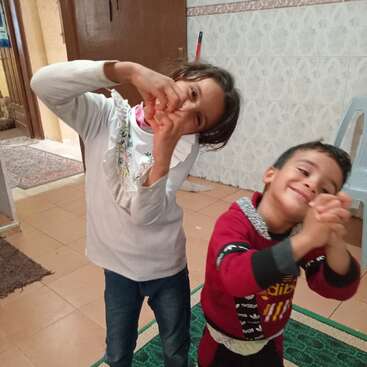 Dos niños alegres están de pie dentro de una casa, sonriendo alegremente. Forman corazones con las manos, mostrándose cariñosos y juguetones, disfrutando juntos de un momento divertido y feliz.