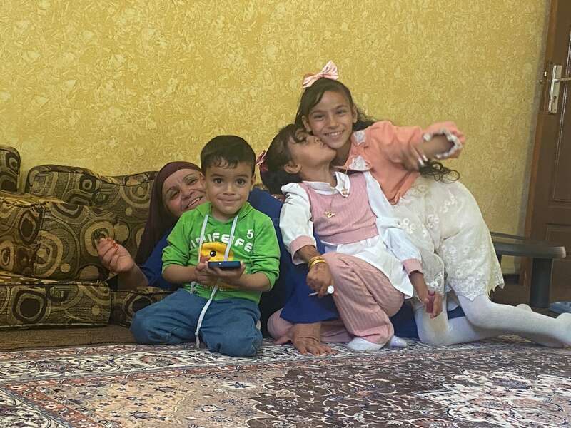 La imagen muestra a una mujer y tres niños sentados en el suelo, la mujer con un pañuelo en la cabeza y los niños vestidos con ropa informal, posando para una foto.