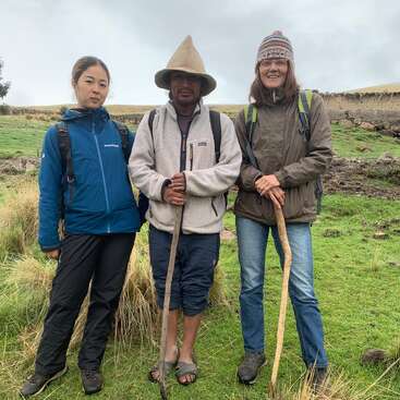 La imagen muestra a tres individuos de pie en un campo cubierto de hierba, cada uno con un bastón en la mano, dos con mochilas y el del medio con un sombrero cónico.