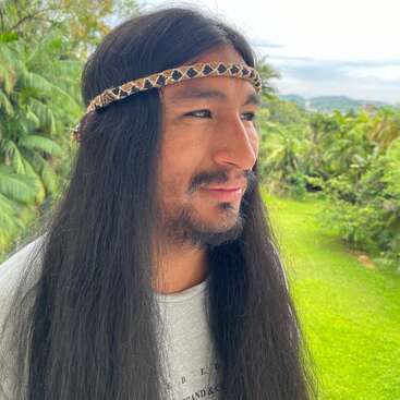 A man with long black hair, wearing a patterned headband, stands outdoors surrounded by lush greenery. He smiles gently and looks to the side, relaxed.