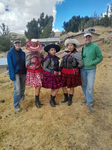 Cinco personas posan al aire libre. Tres mujeres visten ropas y sombreros andinos de colores tradicionales, mientras que dos personas vestidas informalmente con vaqueros y chaquetas sonríen a su lado. Fondo rural.