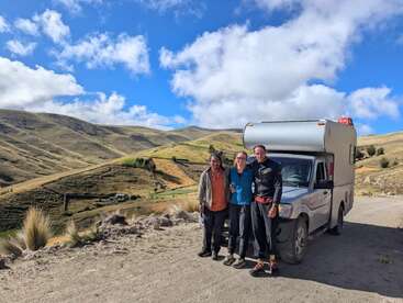 Tres personas sonríen delante de una furgoneta camper en un camino de tierra, rodeado de ondulantes colinas verdes bajo un cielo azul brillante con nubes.