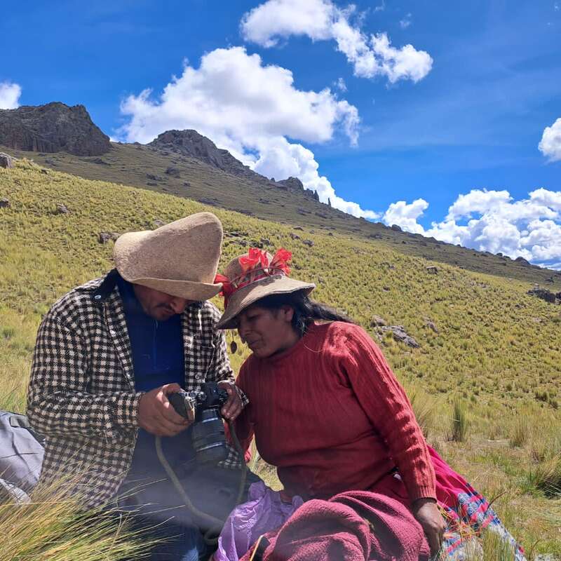 A man and woman in traditional hats examine a camera together, sitting on grassy hills under a bright blue sky with scattered clouds and mountains.