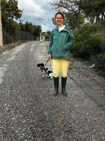 A woman in a green jacket, yellow pants, and green boots stands on a gravel road holding a dog leash, with a small black-and-white dog.