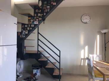 This image shows a modern home interior with a metal staircase decorated with magnets, a clock on the wall, dining table, floor lamp, fridge, and sunlight.