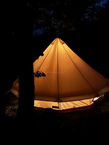 A warmly lit canvas tent glows in the darkness of night, surrounded by trees, creating a cozy and inviting camping atmosphere under the twilight sky.